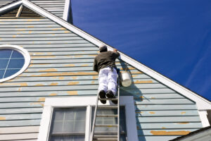 An exterior painter on a ladder applying white paint to the blue siding of a house.