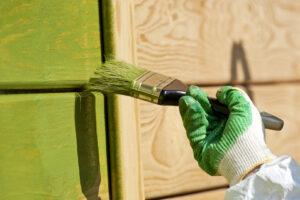 A close-up of a hand holding a green paintbrush, showcasing the work of local exterior house painters as it applies a fresh coat of paint to a wall.