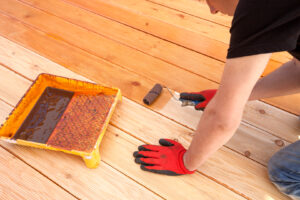 A man applying a dark stain to a wooden deck, working meticulously with a brush. The image captures a sunny day with the focus on the deck staining service in action.