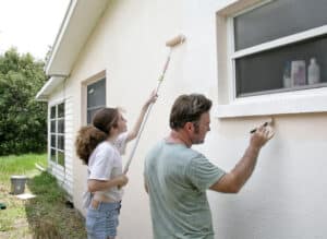 A couple works together as exterior house painters, applying a fresh coat of paint to the siding of a suburban home. The scene captures one person on a ladder while the other mixes paint at the base.