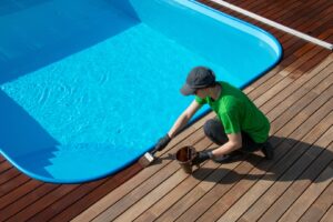 A professional deck stainer using a roller to apply a protective stain on a deck near a swimming pool, ensuring an even coat over the wood.