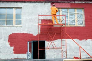 A painter, secured on scaffolding, is covering the exterior of a house with bright red paint, demonstrating skills typical of the best exterior painters.