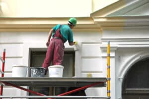 Exterior house painters stand on scaffolding, using a roller to apply paint on a large house. He is focused and methodical in his approach. He wears a safety harness.