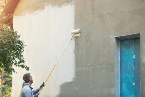 A man, identified as one of the best exterior painters near me, is using a roller to apply white paint to an outdoor wall under a clear sky.