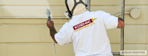 A man wearing a shirt with the Koehn Painting logo is painting the exterior of a house with a roller. The focus is on his back, showing the logo clearly. This image highlights the work of exterior home painters.