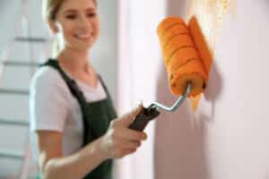 A woman is painting a wall with a roller while two guys observe the process on an outside red wall. This image depicts the activity of commercial building painting.