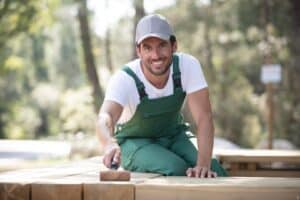 A man is looking into the camera and smiling while painting outside. He is holding a paintbrush and standing near a partially painted wall. This image represents the work of the best exterior painters.