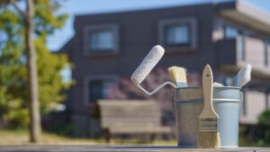 Brushes and rollers are scattered on the grass in front of a house, ready for exterior house painting.