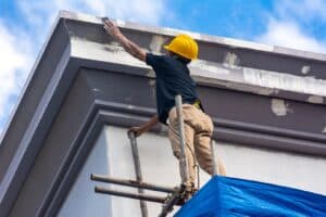 A person is standing on scaffolding and painting outside. They are wearing safety gear and carefully applying paint to the exterior of a building. This image highlights considerations for when not to paint outside.