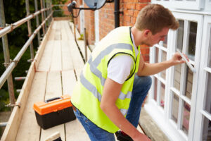 A worker from Koehn Painting is applying paint to the outside walls of a house. He is on a ladder, carefully painting the trim. This image shows professional exterior painters at work.