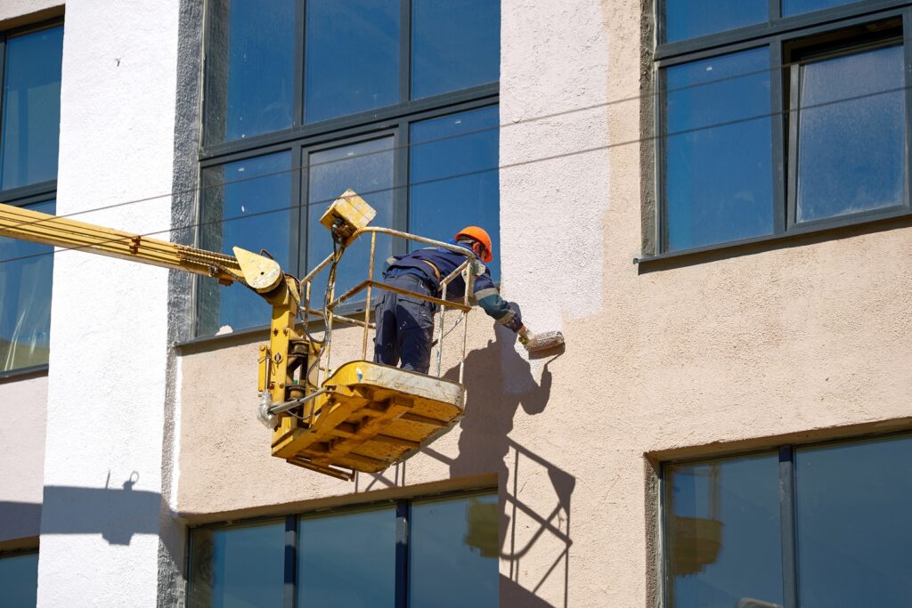 A painter on a lift applying a fresh coat of paint to a commercial building’s exterior, illustrating the scope of commercial painting.