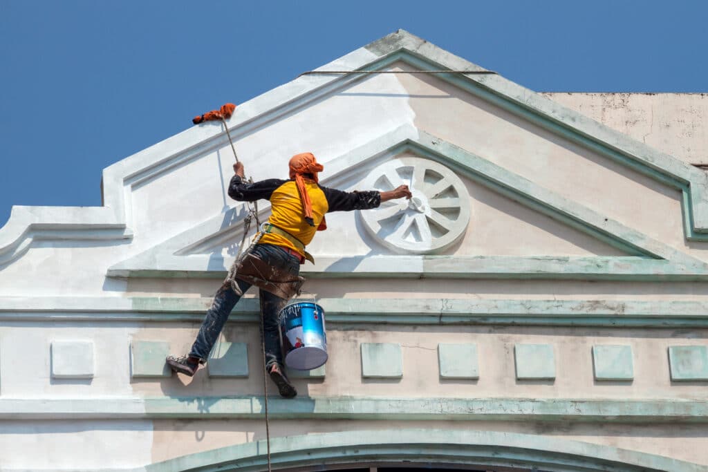 A painter using a rope to access and paint the exterior of a commercial building, representing the work done by a commercial painting company.