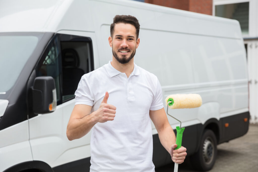 A commercial painter gives a thumbs-up while holding a paint roller in front of a white van, indicating readiness for the job.