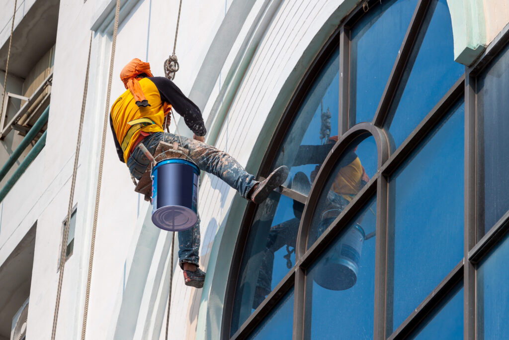 A worker painting a commercial building while suspended on ropes.