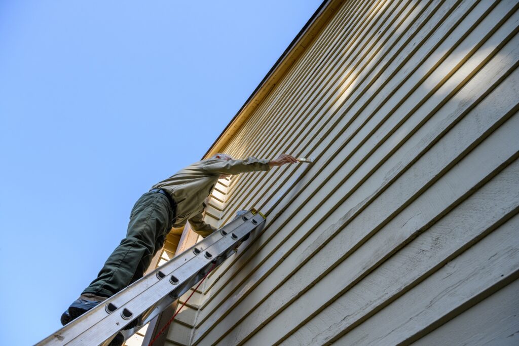 A worker repairing siding on a house using a ladder to apply new paint. The worker is restoring the home's exterior by addressing siding repair with new paint. Regular siding repair prevents extensive home damage.