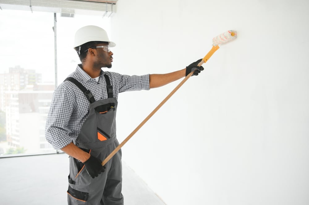 An OSHA-compliant painter, wearing safety gear applies paint to an interior wall using a roller brush.