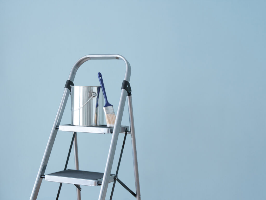 A ladder holding a paint can and brush against a freshly painted blue wall, before a fresh coat of paint. 