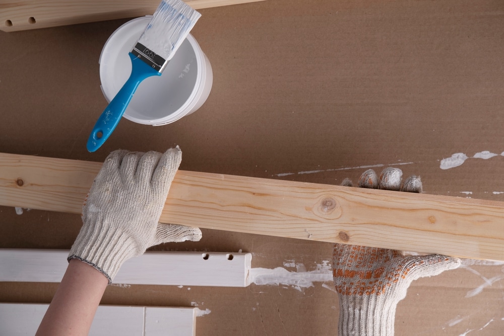 Hands in gloves holding a wooden plank near a paintbrush and white paint, illustrating eco-friendly commercial painting hacks for safe tool cleaning and maintenance