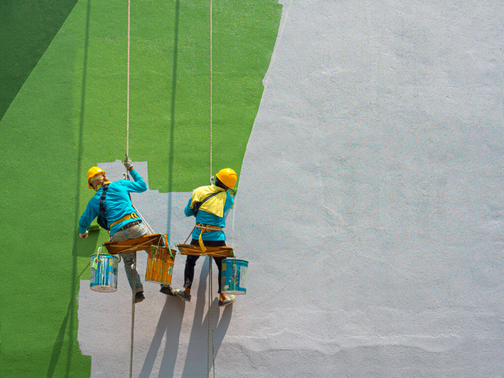 Two professional painters in safety gear working on a green exterior wall, illustrating modern commercial painting trends and techniques in action