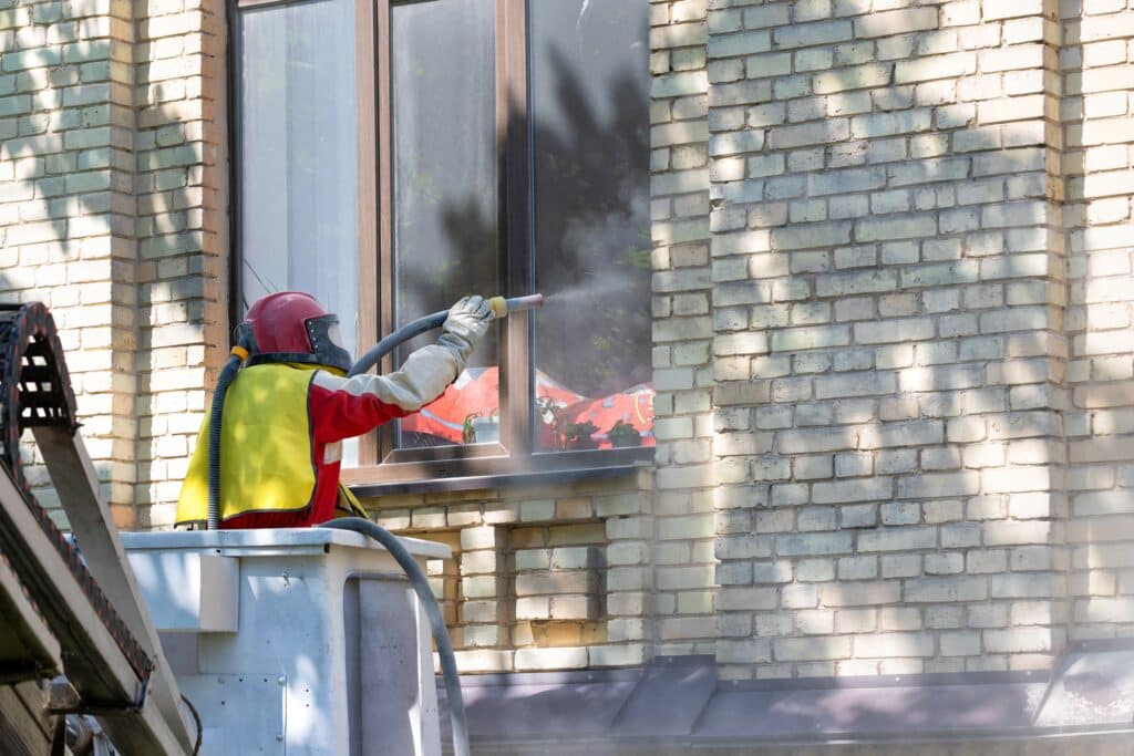 A worker in protective gear performing commercial sandblasting on a brick building exterior.




