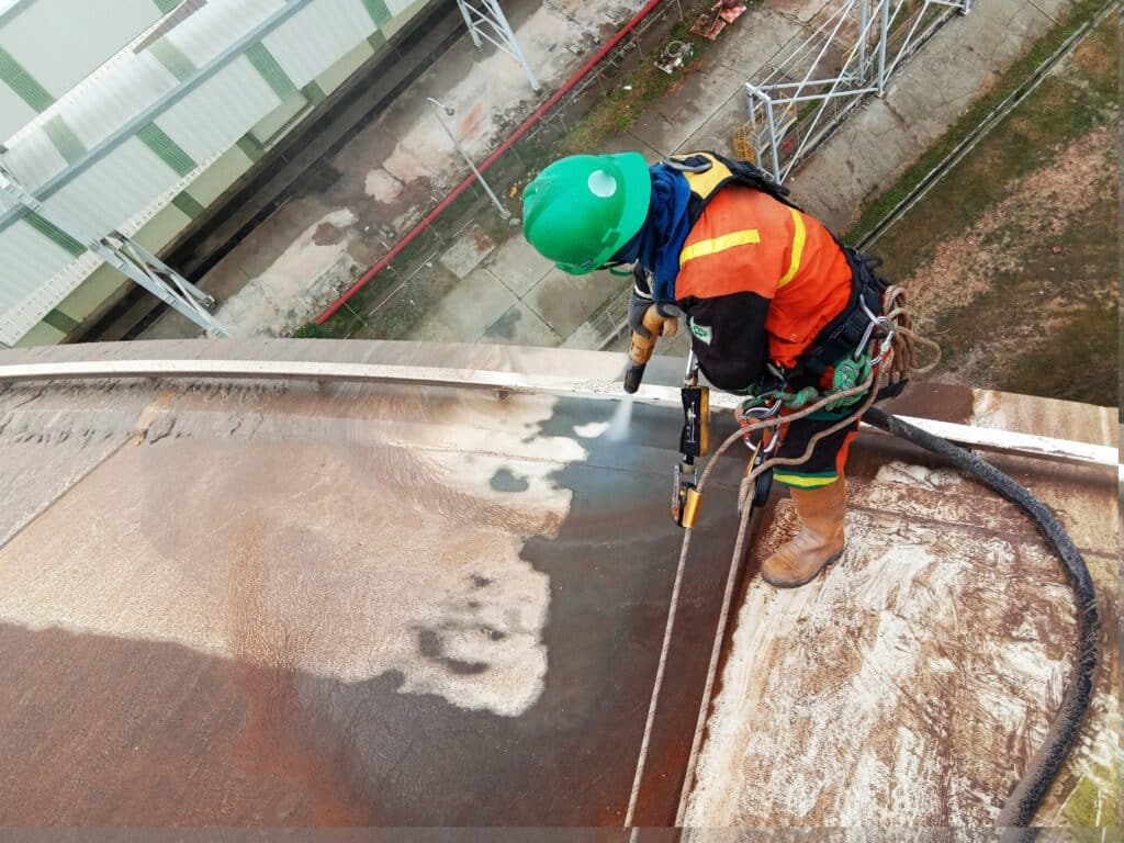 Worker in protective gear performing sandblasting on a large metal surface at an industrial site.




