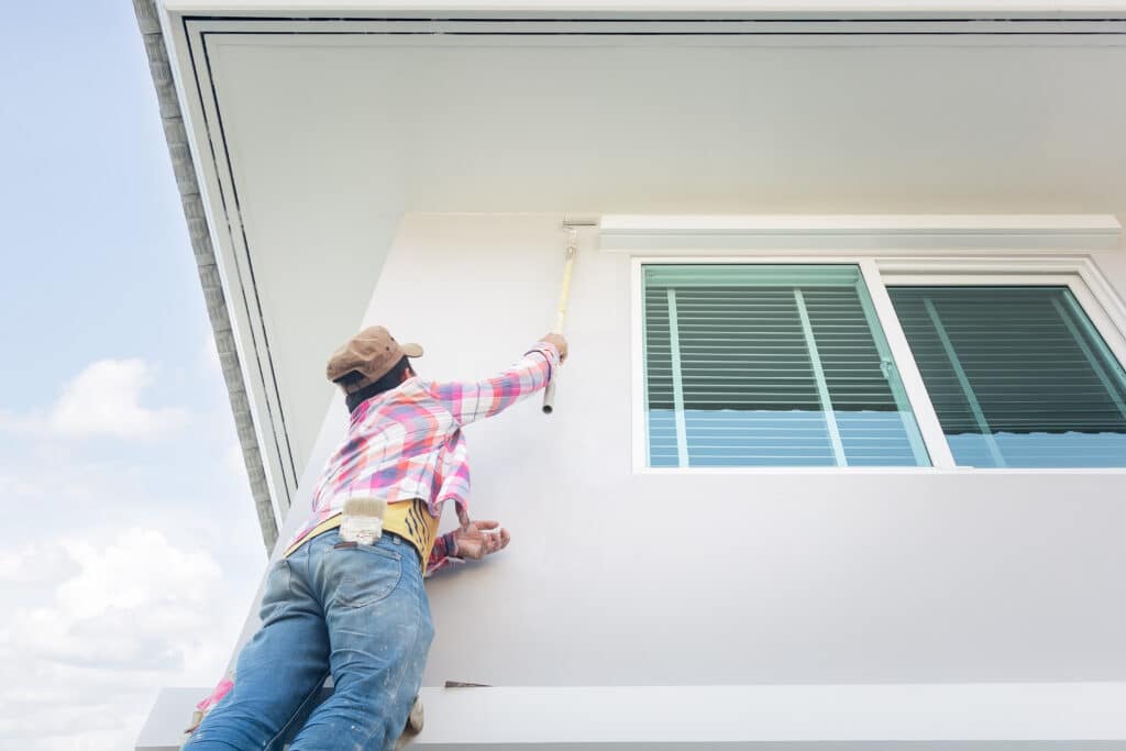 Exterior painters applying fresh white paint to a modern house using a roller brush, reaching high while working near a large window with closed blinds.