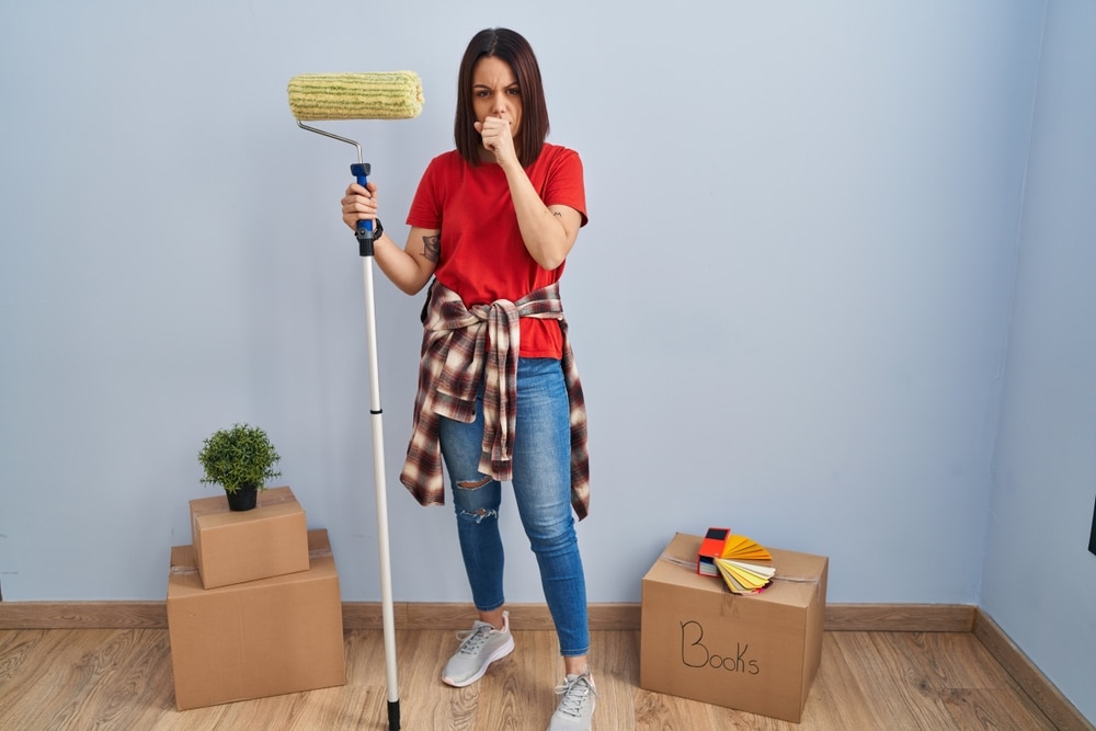 Can you be allergic to paint? A woman holding a roller reacts to paint fumes in a freshly painted room, highlighting potential allergy concerns.