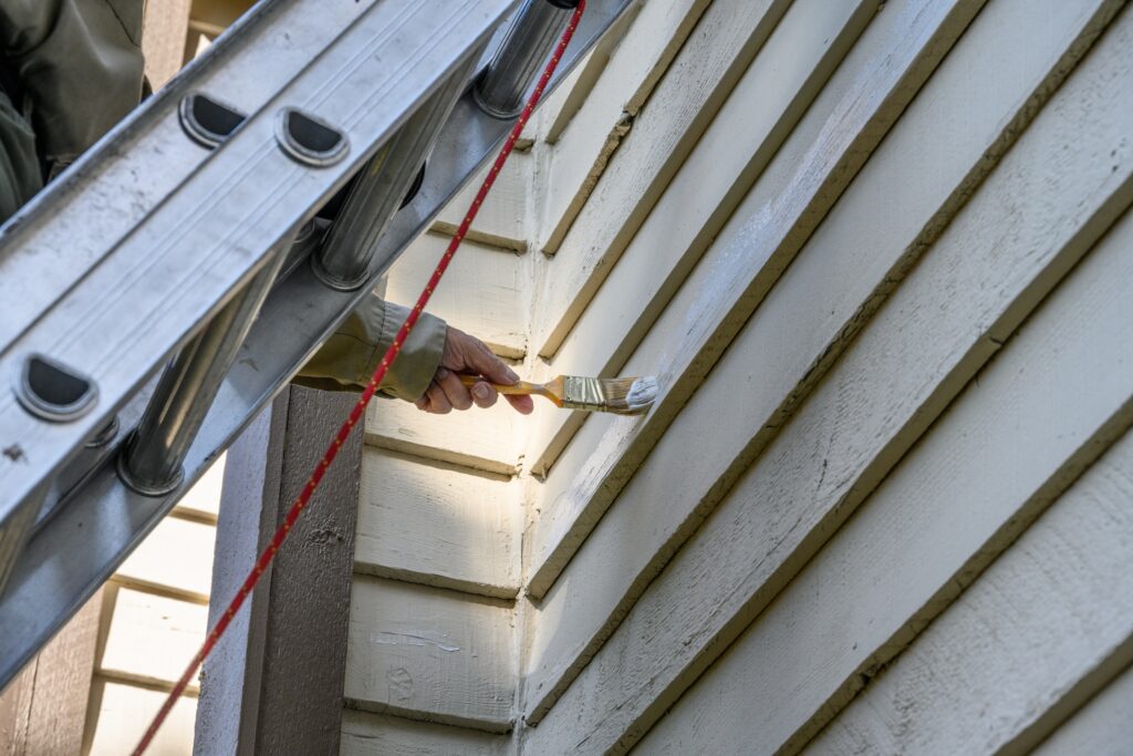 Exterior house painting service in progress! With a professional painter applying fresh white paint to wooden siding using a brush while standing on a ladder.