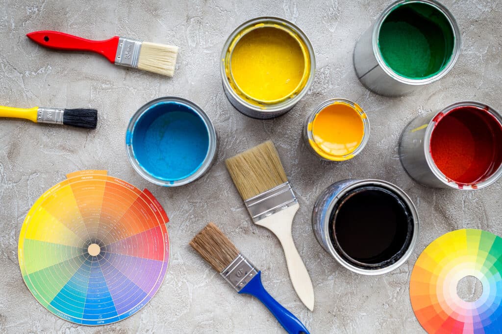 Types of paint finishes displayed alongside paint cans in vibrant colors (yellow, blue, green, red, and orange), brushes, and a color wheel on a textured surface.