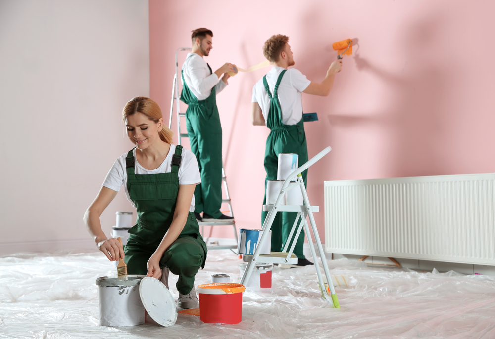 A team of painters in matching green overalls works on a commercial painting project, applying a fresh coat of pink paint to the walls. One painter kneels to prepare a brush with paint, while two others use rollers on the wall, ensuring an even finish. The organized workspace, protective coverings, and high-quality tools highlight the precision and efficiency of a professional commercial painting service.