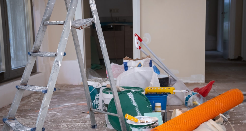 A messy, unorganized painting workspace, filled with scattered paint cans, brushes, rollers, and an old paint-covered ladder. Streaks of paint are visible on the floor, and supplies are left haphazardly around the unfinished walls. This chaotic scene highlights the downside of DIY or unprofessional work—something a skilled commercial painting service could have handled efficiently and cleanly.