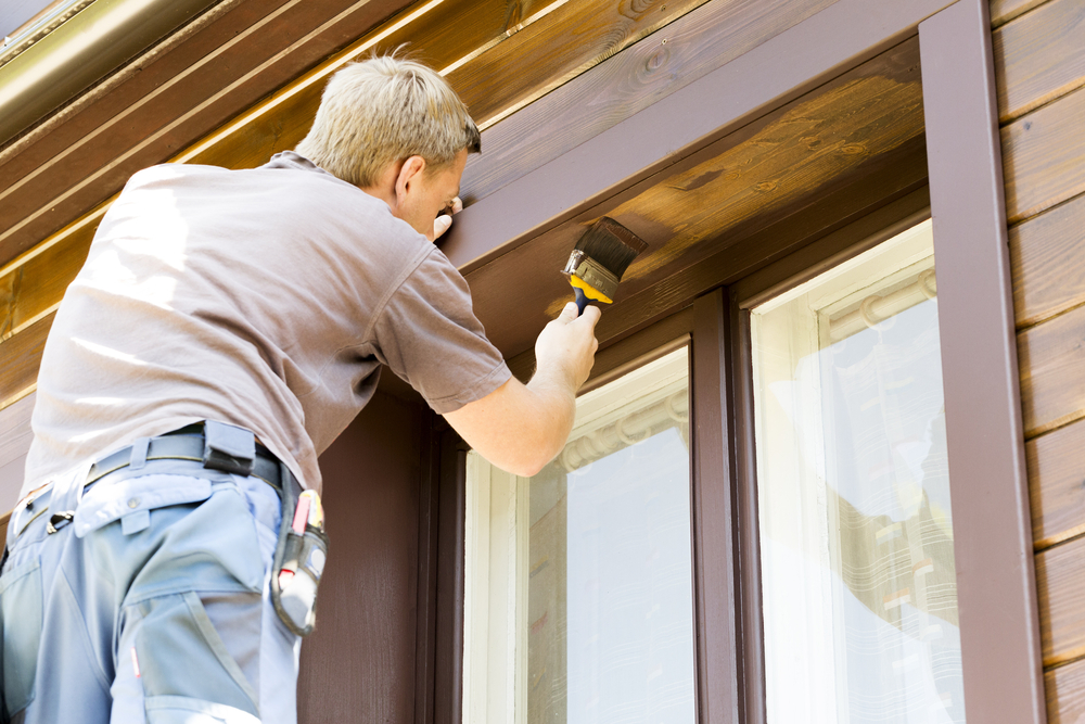 A professional painter carefully applies a fresh coat of brown paint to the exterior wood trim of a house. Equipped with a brush and work gear, he focuses on achieving a smooth and even finish. The detailed craftsmanship highlights the factors influencing exterior painting cost, including labor, materials, and the complexity of the job.