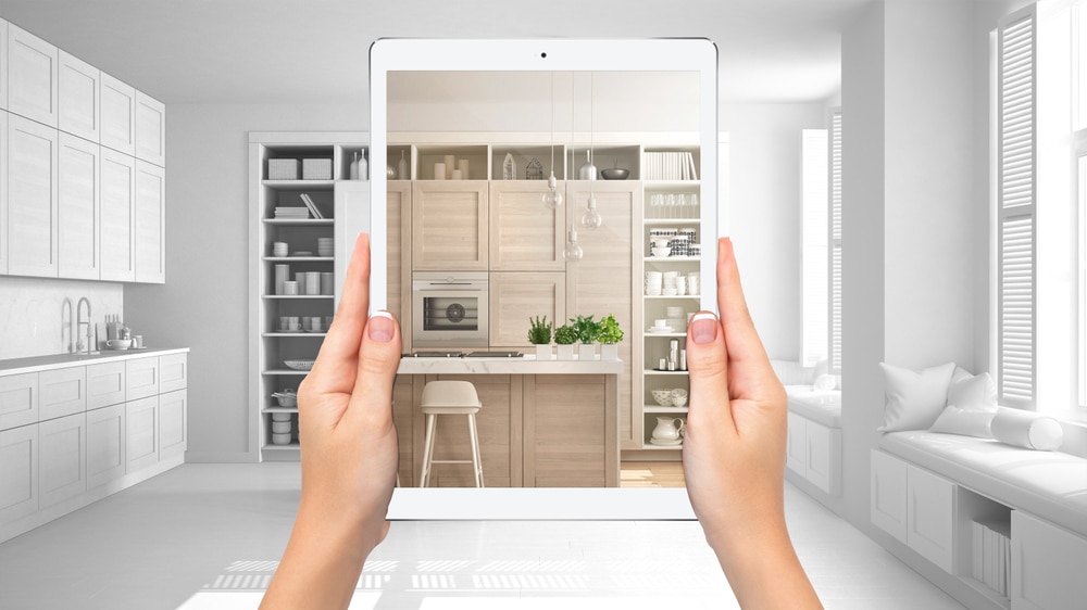 Hands holding a tablet that displays a fully designed modern kitchen over a grayscale sketch background—symbolizing the vision and transformation involved in a total home renovation.