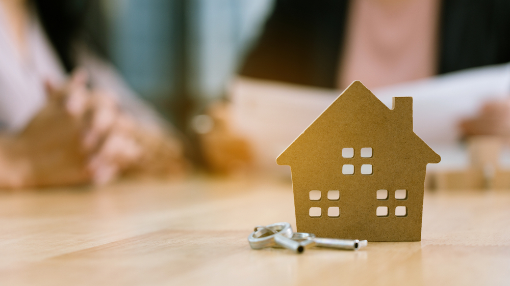 Close-up of house-shaped cutout and keys on a table, with people in the background discussing how to finance a home renovation.