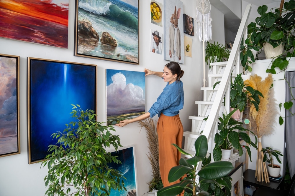 Woman arranging artwork on a gallery-style wall inside a vibrant, plant-filled home, showcasing unique renovations that blend art, nature, and personal expression.
