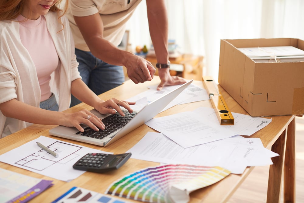 Couple reviewing renovation plans on a laptop surrounded by color swatches, blueprints, and a calculator, representing a hands-on approach to 'how to budget for a home renovation' by planning expenses and making informed design choices.